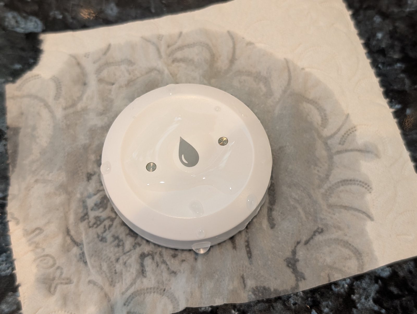A leak sensor sitting on kitchen tissue so small drips can spread toward the contacts